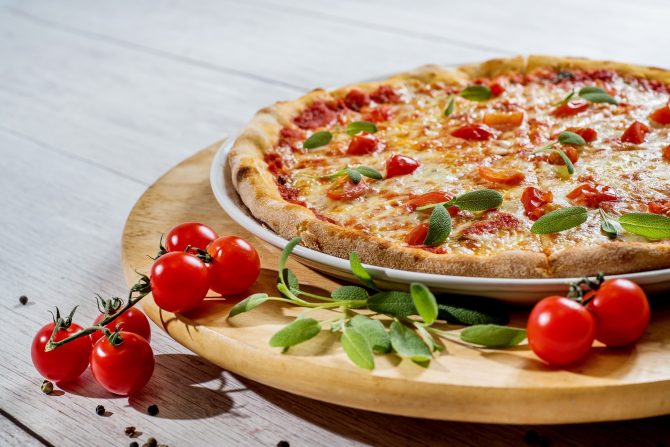 Pizza na drvenom pladnju koji se nalazi na stolu okružen cherry rajčicama i bosiljkom